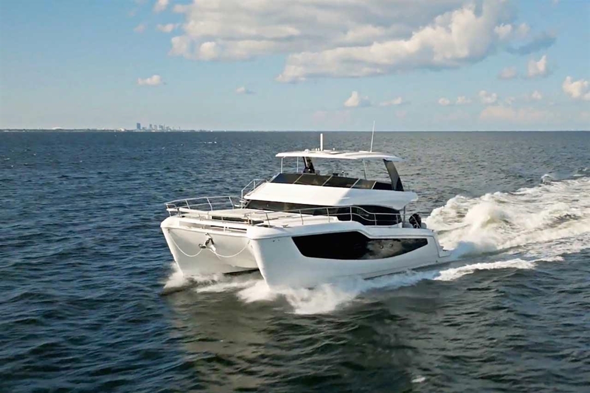 A luxury catamaran sailing on calm waters, leaving a wake, with a skyline visible in the distance under a blue sky.